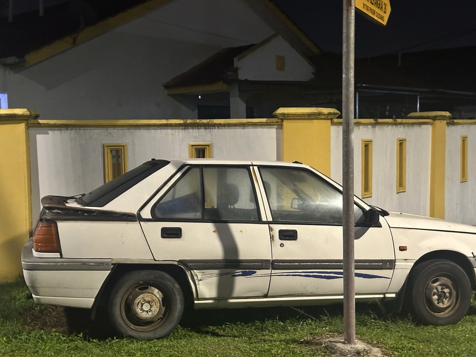 Old Proton Saga parked next to yellow-walled building — dented, faded, still running