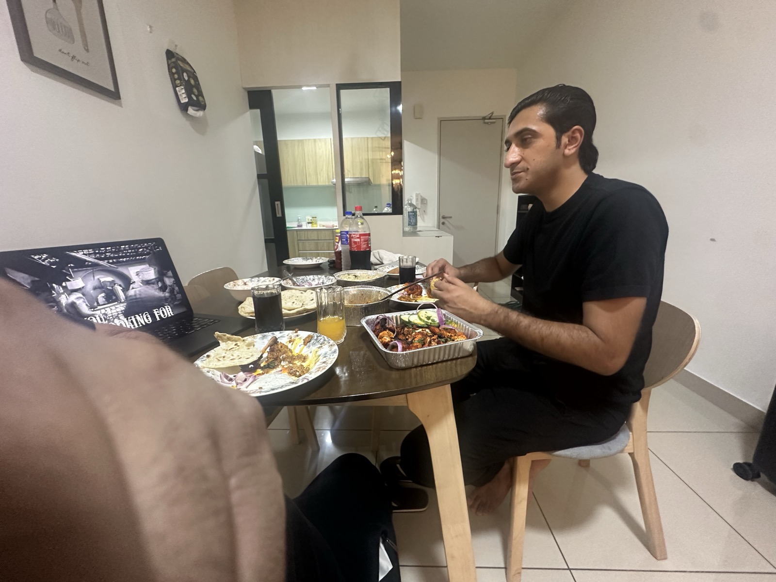 Apartment dinner in JB — Haadi eating, Pakistani takeout spread, MacBook showing 'YOU ARE LOOKING FOR ADVENTURE' — Day 5