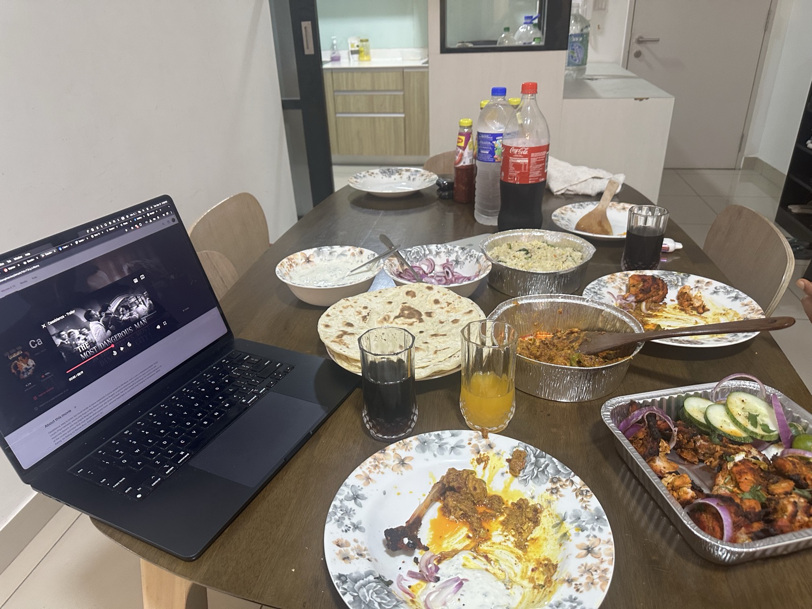 Apartment dinner wide shot — full spread: biryani, karahi, naan, Coca-Cola, MacBook showing 'THE MOST DANGEROUS MAN'