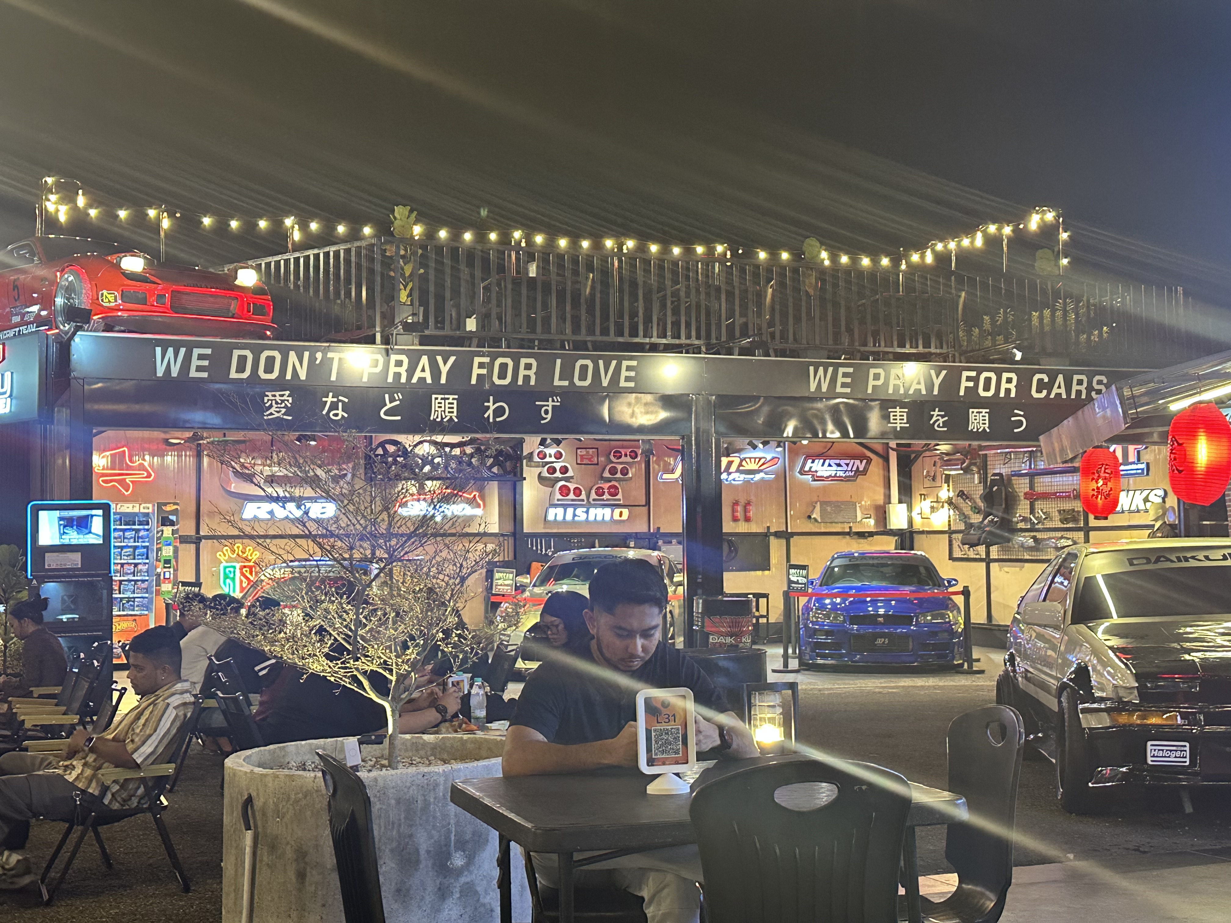 The J — WE DON'T PRAY FOR LOVE WE PRAY FOR CARS, red car on roof, blue Skyline, Nismo signs, string lights