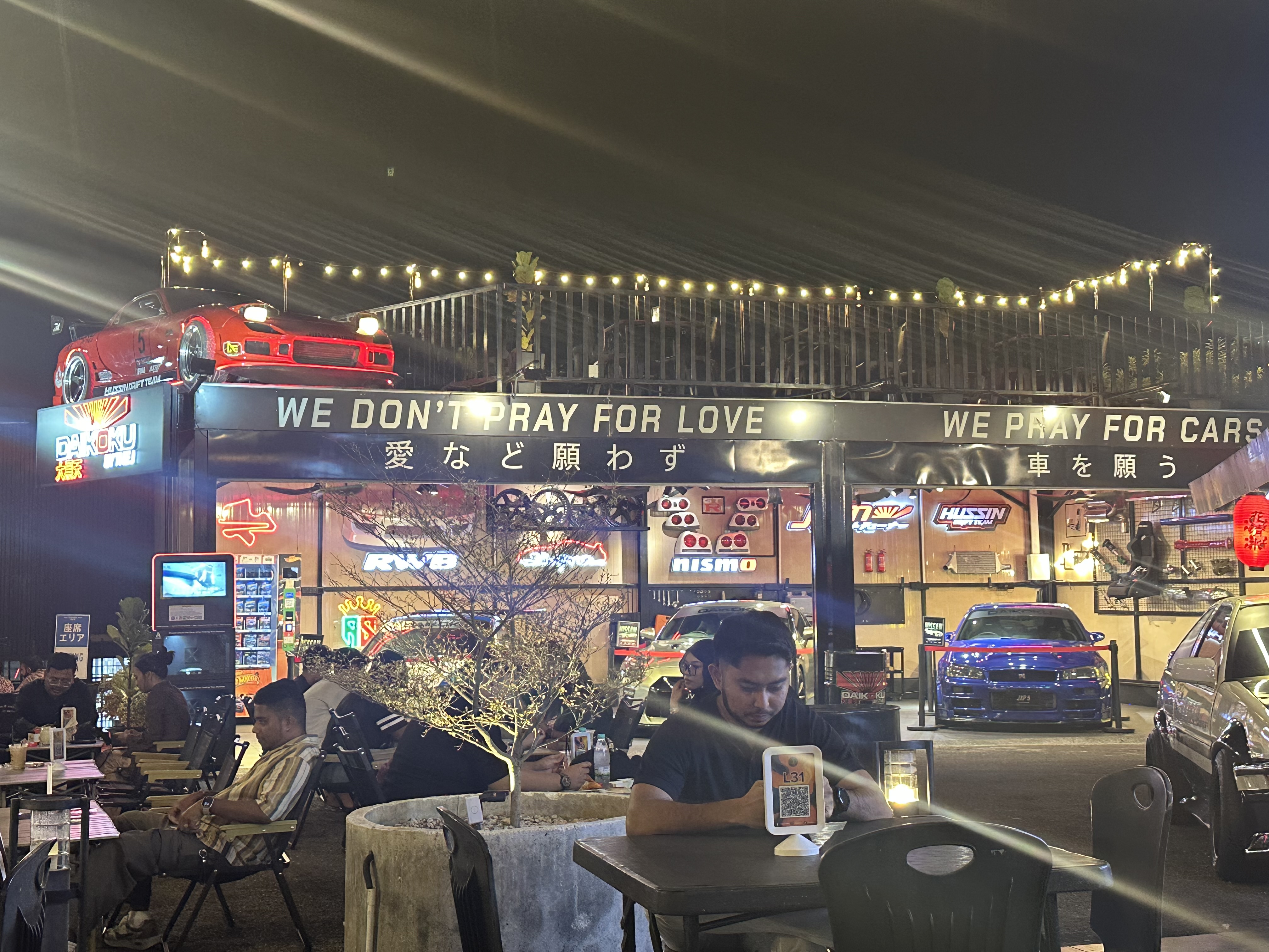 The J wide shot — full car-culture venue with tuned cars, diners, neon