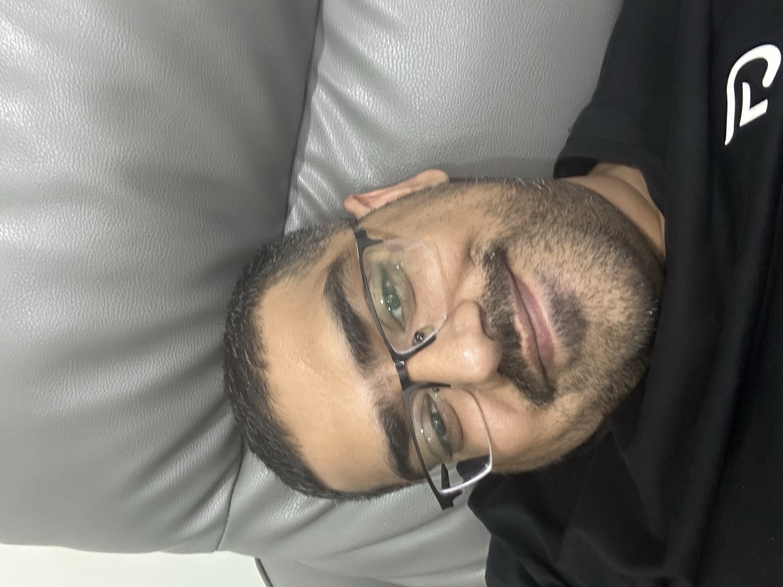 Omer selfie on leather couch — black shirt, glasses, hotel room in Johor Bahru